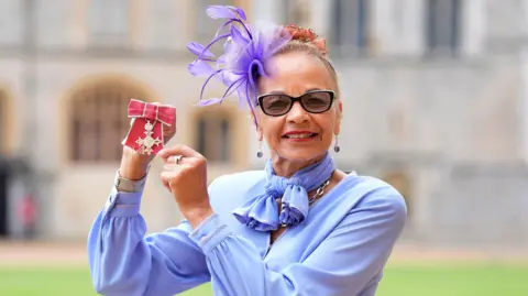 PA Media Anita Neil, who is wearing a purple blouse, scarf and fascinator, smiles as she holds up her award in the grounds of Windsor Castle