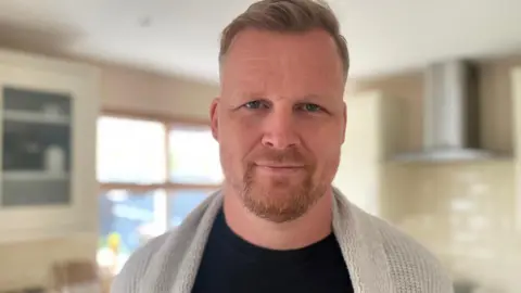 Gemma Dawson/BBC Head and shoulders shot of Joe Barker. He has light-coloured slicked back hair and a short beard. He is wearing a black top and a beige cardigan.