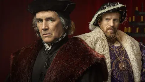 A promotional image for the Wolf Hall series. To the right is Mark Rylance as Thomas Cromwell in period costume with a black hat and top and a claret fur cloak. To the left is Damian Lewis dressed as King Henry VIII with a feathered hat, white cape and purple tunic.