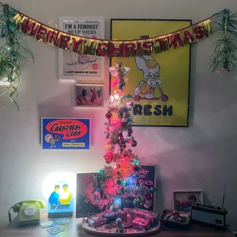 Lucy Scott A small Christmas tree on a table, with baubles and an angel, with some posters in the background and a banner that says "Merry Christmas" above it