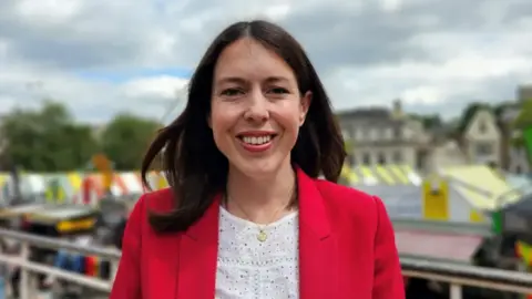 Paul Moseley/BBC Profile picture of Alice MacDonald pictured outside Norwich Market, smiling at the camera. She has long brunette hair, red lipstick, a gold necklace, and a white top with a red suit jacket over the top. 