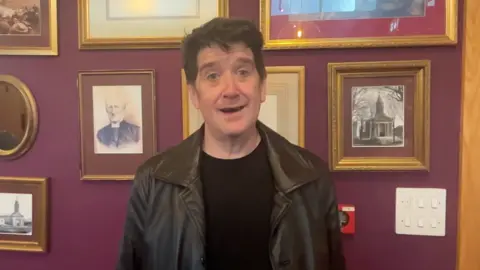 A smiling man with dark hair, wears a leather jacket and black t-shirt in front of a purple wall filled with old photos and drawings of people and buildings from Guernsey.