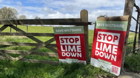 Two posters mainly coloured red saying "SAVE WILTSHIRE STOP LIME DOWN" are rested against a gate to a field. In the field behind can be seen grass and trees. 