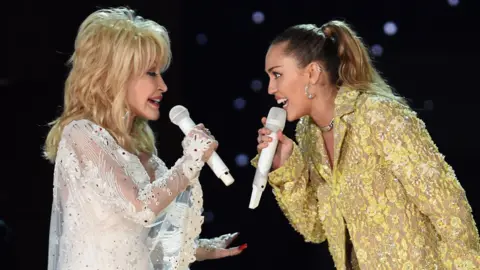 Dolly Parton and Miley Cyrus facing each other, both singing into microphones on stage