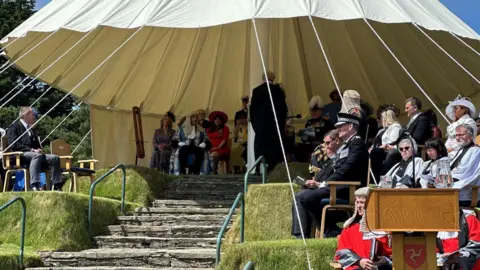 Dignitaries seated on Tynwald Hill in St John's, which is a tiered grass hill with a canopy over the top.