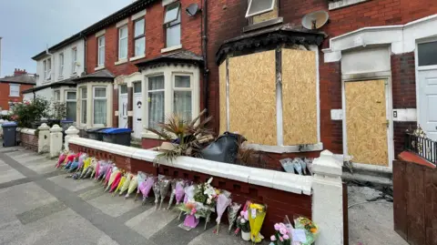 Floral tributes left outside a house gutted by a fire in Blackpool. The windows and door of the home is boarded up. Flowers with different coloured wrappings are lined up against a small brick wall on the public pavement.