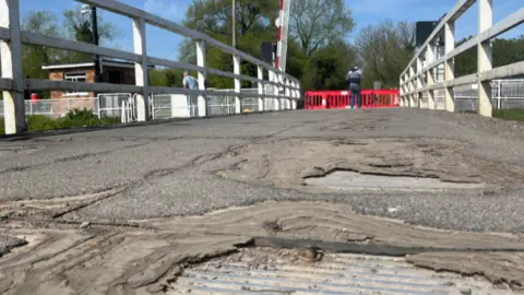 An uneven road surface on the swing bridge. There is a red gate at the end of the bridge, blocking it off. Two men are stood at the end of the bridge. There are trees in the background. It's a sunny day and the sky is blue.