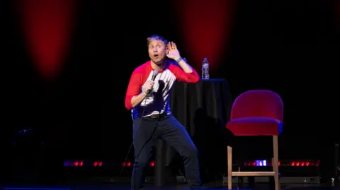 Craig Sugden Blonde man in black trousers and a red and white t-shirt on stage holding a microphone. He has his mouth open and his hand cupped to his ear - and looks a bit silly.