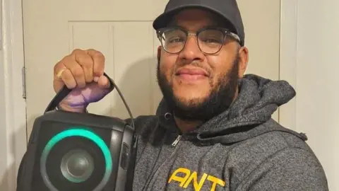 supplied A man in a dark baseball cap and grey hoodie holds up a large black speaker to the camera