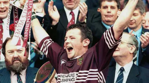 Richie Gray A dark-haired man wearing a maroon rugby top holds up a trophy, with many people in the background looking on
