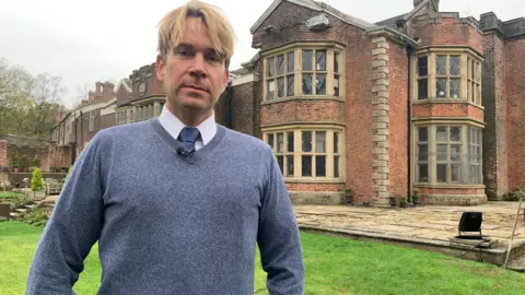 BBC Hopwood DePree stands outside Hopwood Hall, in Middleton, Rochdale. He has blond hair and his wearing a tie under a blue jumper. 