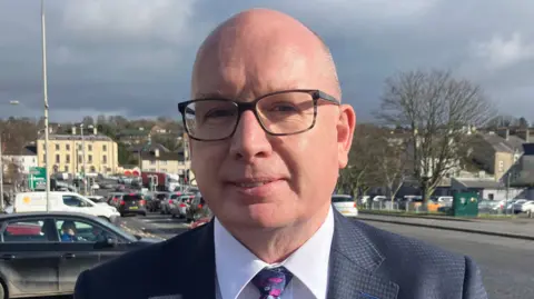 Mr Savage is bald and is wearing rectangular glasses, he has a white shirt and is wearing a black suit with a patterned tie. Behind him is a street where cars are parked along the side and queued up at a set of traffic lights. In the background there are buildings.