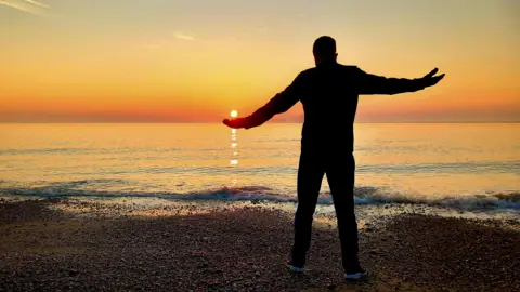 Hayley85/BBC Weather Watchers A man stands on a beach during sunset. He has placed his hand slightly below him so it looks like he is holding the sun in his palm. 