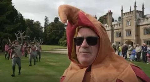 Terry Bailey wearing a red and orange jester costume with dancers behind him carrying the deer antlers. Behind him on the right is Blithfield Hall, one of the venues the dancers visited. 