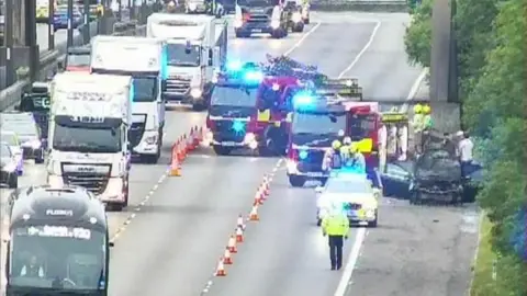 National Highways A motorway, with traffic cones on it and two fire engines, a burned out vehicle to the left, and cars travelling past a number of cones. Emergency workers are walking on the road. There are queues of traffic. 