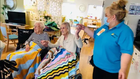 Two elderly women using wheelchairs smiling and holding hands with a younger woman in a blue polo short.