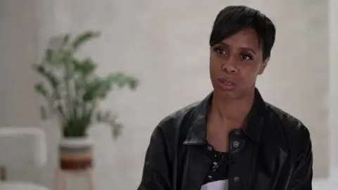 Serlina Boyd has short black hair in a pixie cut. She is wearing a black leather shirt and a sequin dress. She is sitting in a room against a beige backdrop with a plant in the distance behind her.