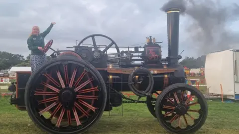 CLAIRE WHITEWAY Claire on a large steam traction engine at the show. She has her left hand raised while her other is on the steering wheel.