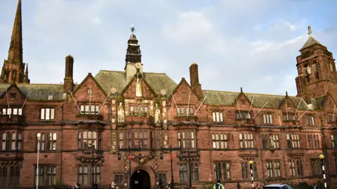 A building of at least three storeys, with an arched entrance at the bottom. There is a spire on the far left and the blue sky with white clouds is visible above the building.