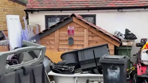 Tall piles of rubbish, including a bath, wheelie bins, a door and seating ripped out of a van. It is all laid out in the backyard of Save A Paw. There is also a small shed with a red sign that reads: "No trespassing. We're tired of hiding the bodies".