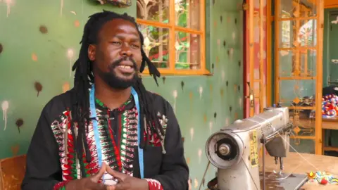 BBC Fashion designer Latif Madoi sits next to a sewing machine