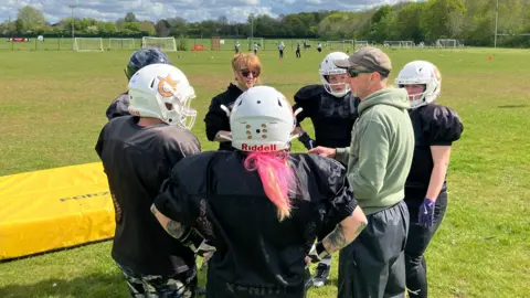 Shariqua Ahmed/BBC Players standing in a huddle - wearing white helmets, with coach Maurice,  man, standing in middle talking to them.