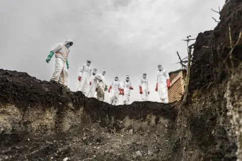 MICHEL LUNANGA / AFP Undertakers line up at the edge of a dug mass grave. It is empty, ready for bodies to be laid to rest in it.