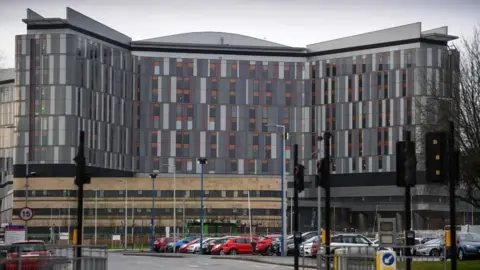 Getty Images Edifício principal do Hospital Universitário Queen Elizabeth, tirado da rua, com semáforos e carros estacionados em primeiro plano.