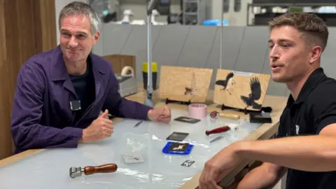 BBC/Lucinda Adam Peter Kyle sits at a work table opposite a Rolls Royce employee. There are tools on the table between them and examples of detailed artwork from car interiors.