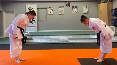 A mother and daughter, wearing their black karate belts, bow to one another before sparring.