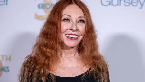 Getty Images Cassandra Peterson, smiling at an event in California, wearing a black top and a white backdrop.