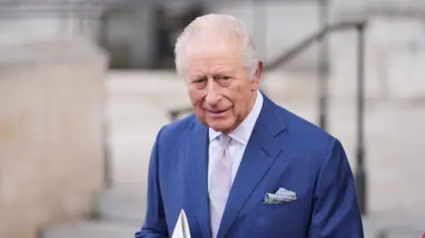 PA Media An elderly man in a blue suit, white shirt and mauve tie smiles outside a building.