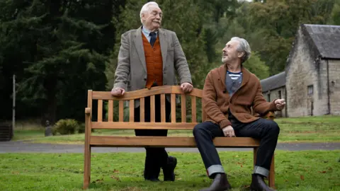 Lisa Birch Alan Cumming, in jeans and a brown cardigan, sits on a wooden bench, turning to look up to Brian Cox, standing behind the seat. There is a beautiful row of large trees behind them, and a stone building. Cox smiles down at Cumming.