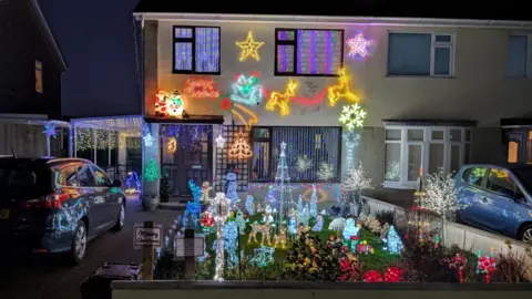 Xanthe Anderson Christmas display in Frome. The house is covered in lights such as stars and a sleigh. The front garden features other light figures like trees, reindeers and penguins. 