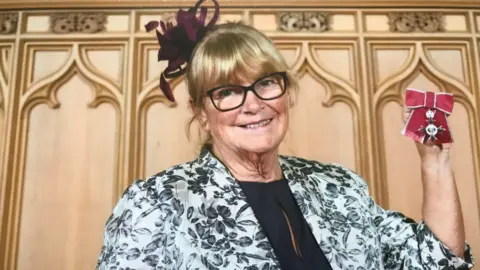 Patricia Rogers A woman with a blonde fringe is wearing a black top and a white floral jacket. She is holding up her MBE medal with its red ribbon.