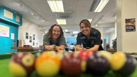 Two women, one of whom wears a police uniform, stand at the end of a pool table and smile at the camera. The pool balls are close to the camera but have been blurred to focus on the two women.