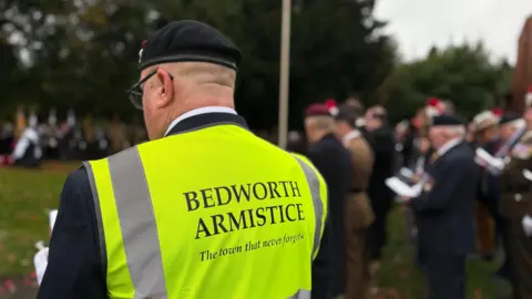 A man is wearing a military beret and a hi-vis vest with the words 'Bedworth Armistice - the town that never forgets' in text on the back. In the blurred background, an Armistice day service is in progress.