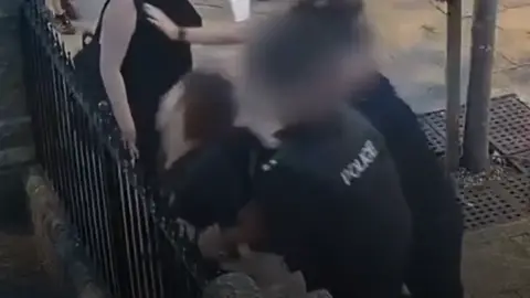 Supplied Slightly blurry CCTV footage showing two police officers behind a young man whom they are trying to restrain up against a metal fence. His head is close to the spikes on the fence. 