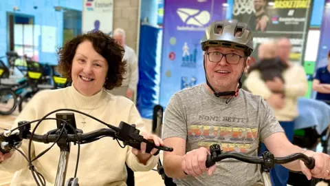 Tony Deeney sits on a dual side by side tandem bike with his wife, Teresa. Tony holds onto the handlebars wearing a grey t-shirt with a grey helmet. He is smiling while looking at the camera. Teresa has short brown hair wears a cream jumper and holds onto the handlebars. She is smiling at the camera. 