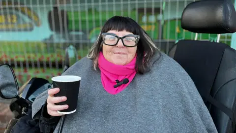 A woman in a grey coat and pink scarf sits in a mobility scooter with a cup of coffee