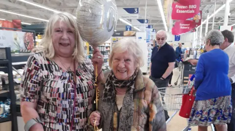 Supplied Lynda Graves, who is standing on the left and holding up a balloon, is next to her 101-year-old mother, Sally Wardle. They are both standing inside a supermarket next to the checkouts. 