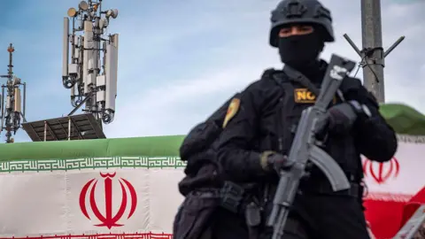 An armed Special Units police officer in Iran, dressed in black and holding a weapon, is standing on an armored vehicle while monitoring the surroundings. In the background, telecommunications towers and the Iranian flag can be seen. The photo was taken during a pro-government gathering in central Tehran.
