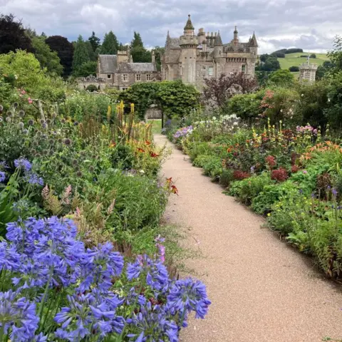 Janice Taylor Path flanked by bright flowers, leading to a grand house in the distance, and countryside behind it.