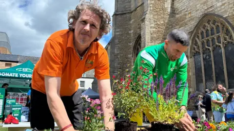 Jenny Kirk/BBC Dave Poulton and David James planting colourful flowers in to a planter outside a church in Peterborough city centre. 