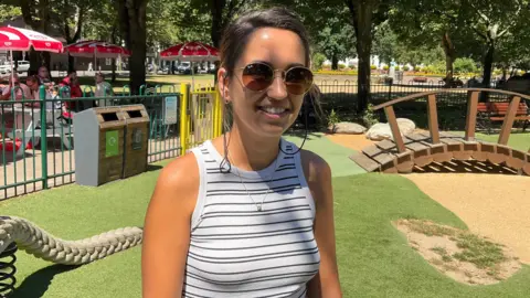 A woman wearing sunglasses smiles at the camera while standing beside play equipment in a play park.