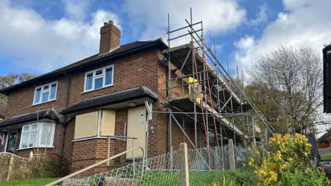 A semi-detached terraced house with boarded up ground floor windows and scaffolding on its right wall