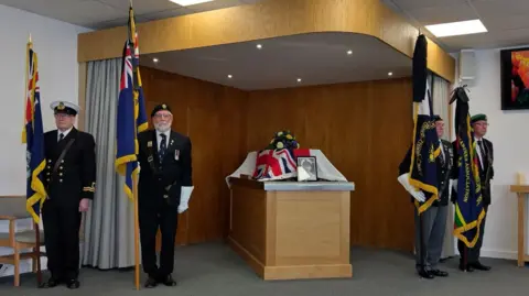Jackie Kirk Four standard bearers stand to attention as they hold their flag poles upright. They wear military uniform and stand two either side of a coffin on a plinth, which is draped with a Union Jack flag. There is a photo of the man as a young sailor at the front of the coffin and a bouquet of flowers on top.