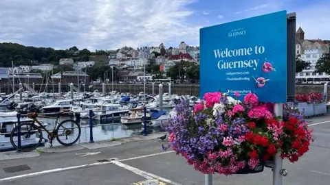 A blue Welcome to Guernsey sign by Guernsey's harbour. It has baskets of flowers beneath it.