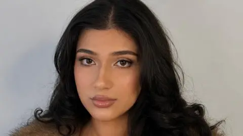 Iqra Wasim Close up of Wasim who has long hair and brown eyes. She is stood in front of a grey background.