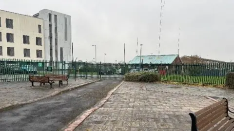 Neighbourhood Network An outdoor paved area with benches and a narrow path lined by red bricks. A green metal fence surrounds the space, separating it from nearby buildings, which include a white structure and a smaller blue-roofed building. The sky is overcast and the pavement is wet.